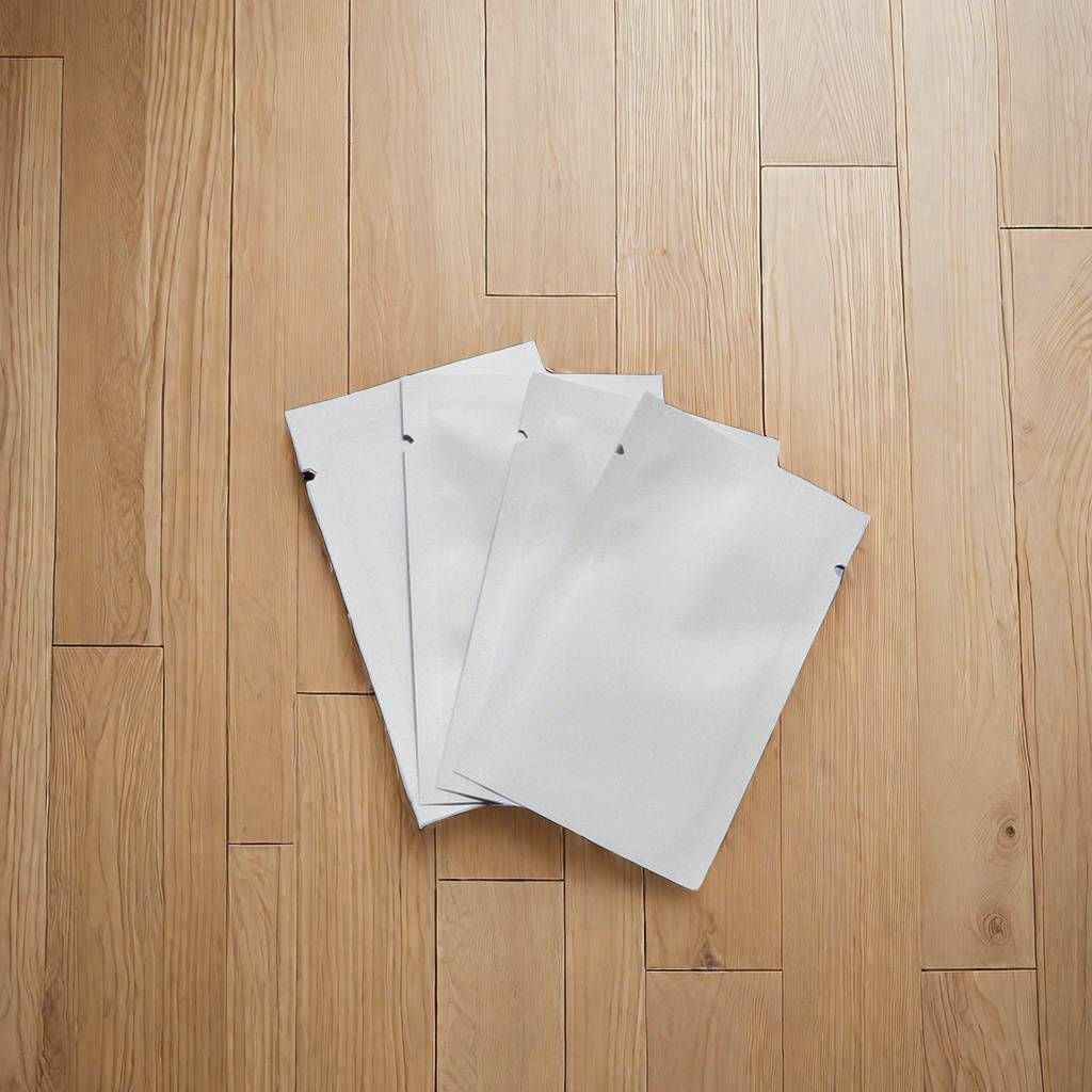 四個空白的白色長方形鋁箔包裝袋層層疊放，擺放在淺棕色的木地板上。
