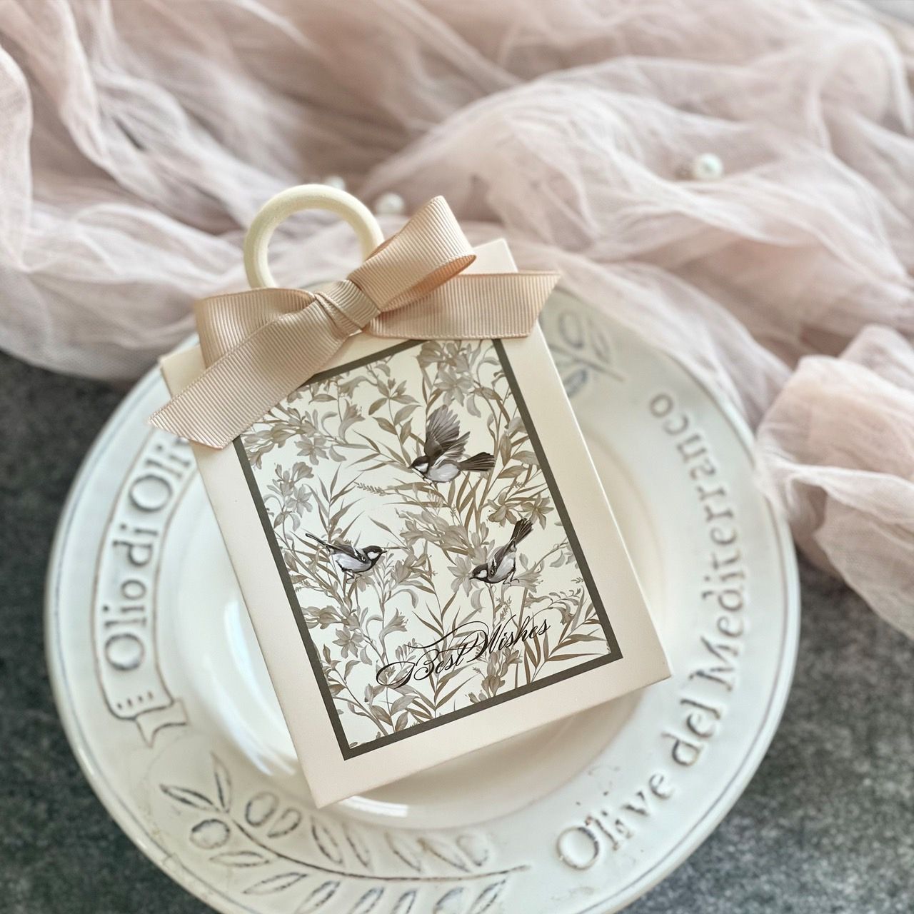 一個米色禮盒，印有鳥類圖案，繫著棕褐色絲帶蝴蝶結，放在一個白色華麗的裝飾盤上。