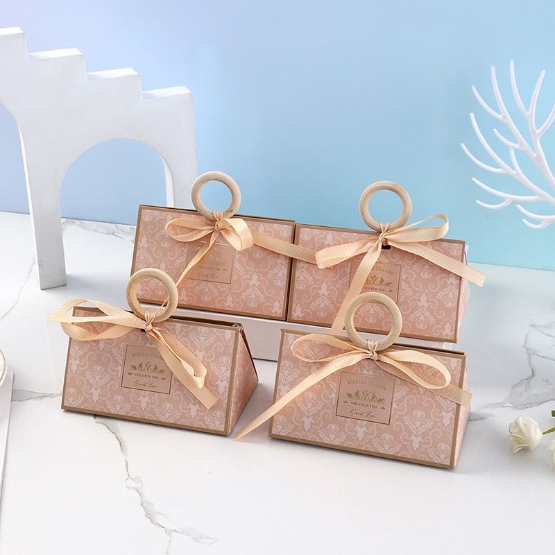 四個優雅的淡粉色禮盒，飾有幾何圖案，配有木質環形提手和米色絲帶。