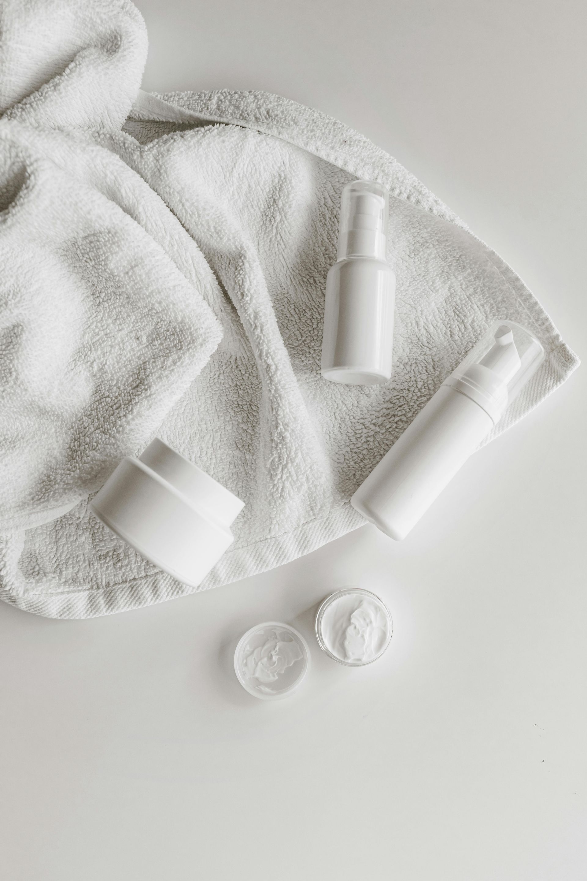 一條白色毛巾、兩個白色護膚品按壓瓶和一個有兩個敞開蓋子的罐子，擺放在純白色的表面上。