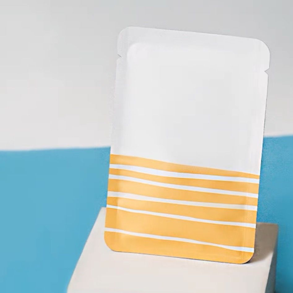 一個長方形的白色護膚品包裝袋，底部有手繪的水平黃色條紋，放在一個底座上。