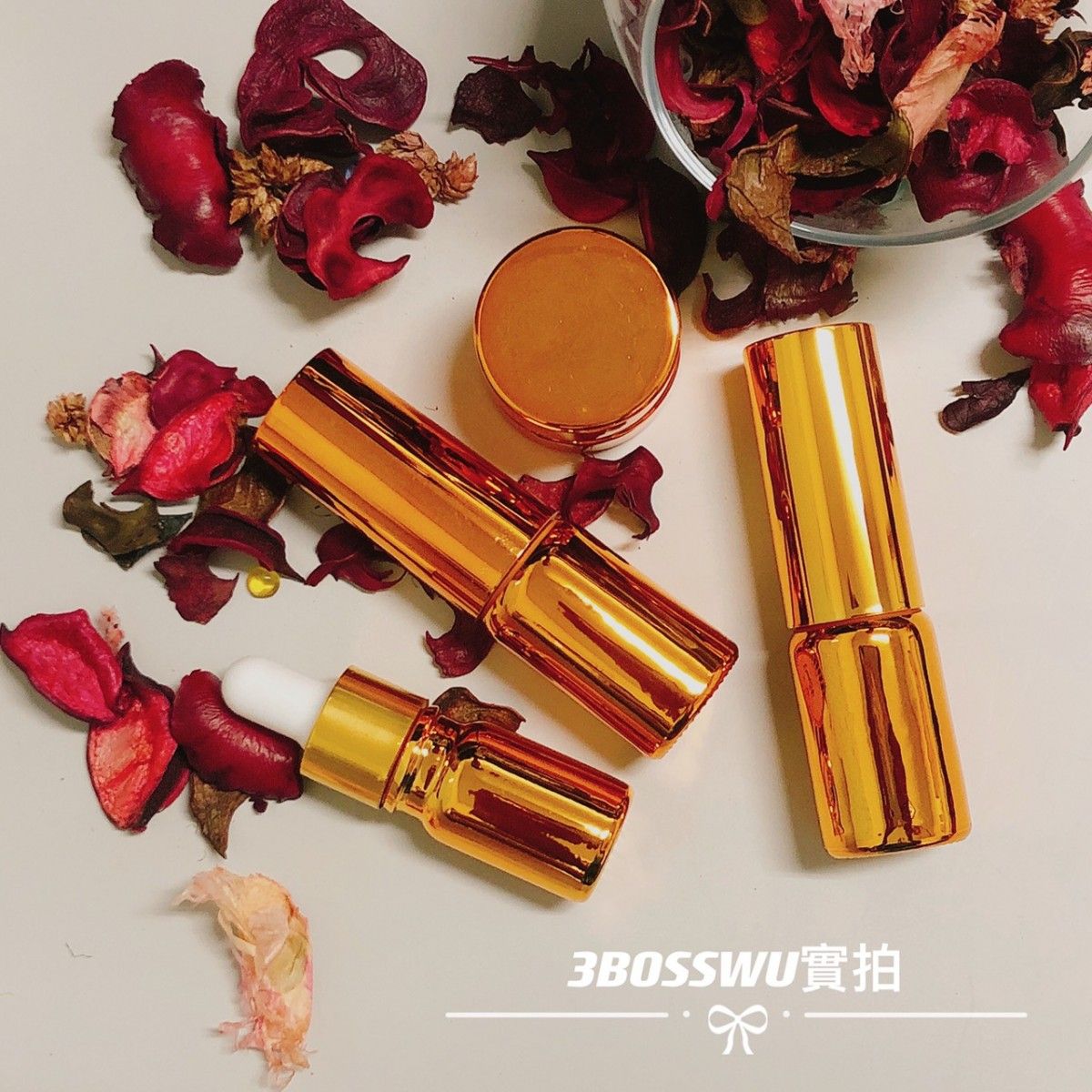 三個金色的化妝品瓶和一個小罐子擺放在一個桌面上，桌面上散落著乾枯的紅色花瓣。