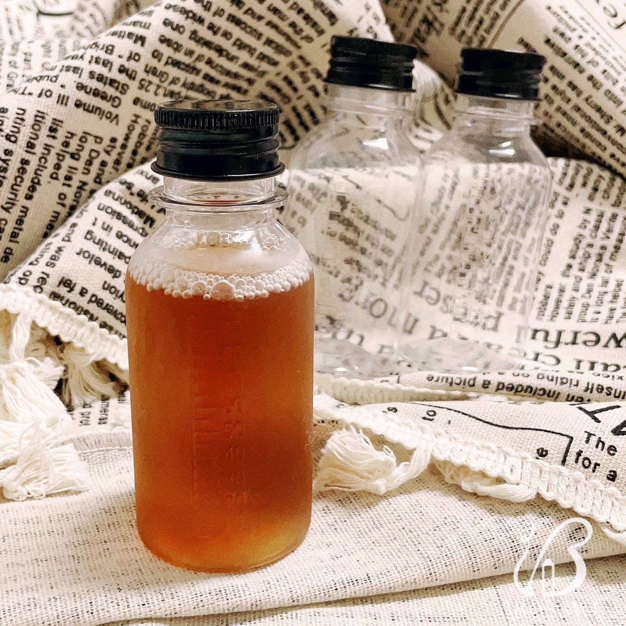 一瓶裝滿琥珀色液體的瓶子擺放在報紙圖案的布料背景上，前面是兩個空瓶。