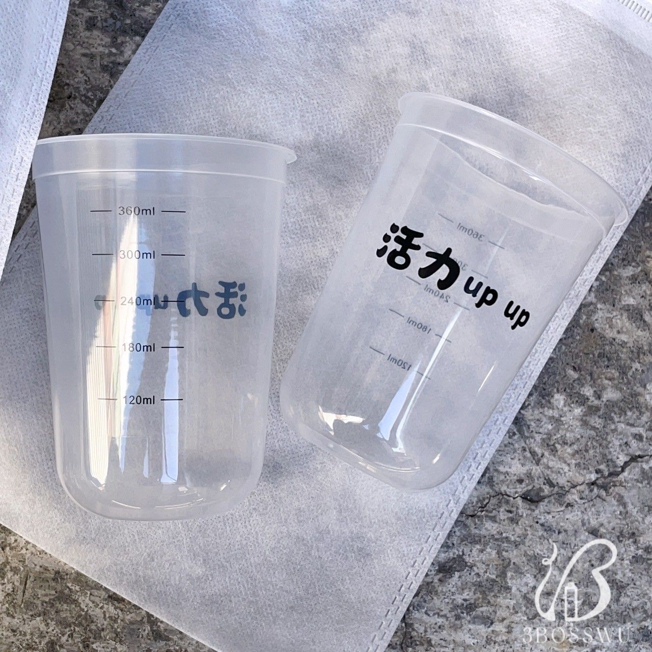兩個透明塑膠量杯，印有黑色文字和刻度線，放置在有紋理的白色織物表面上。