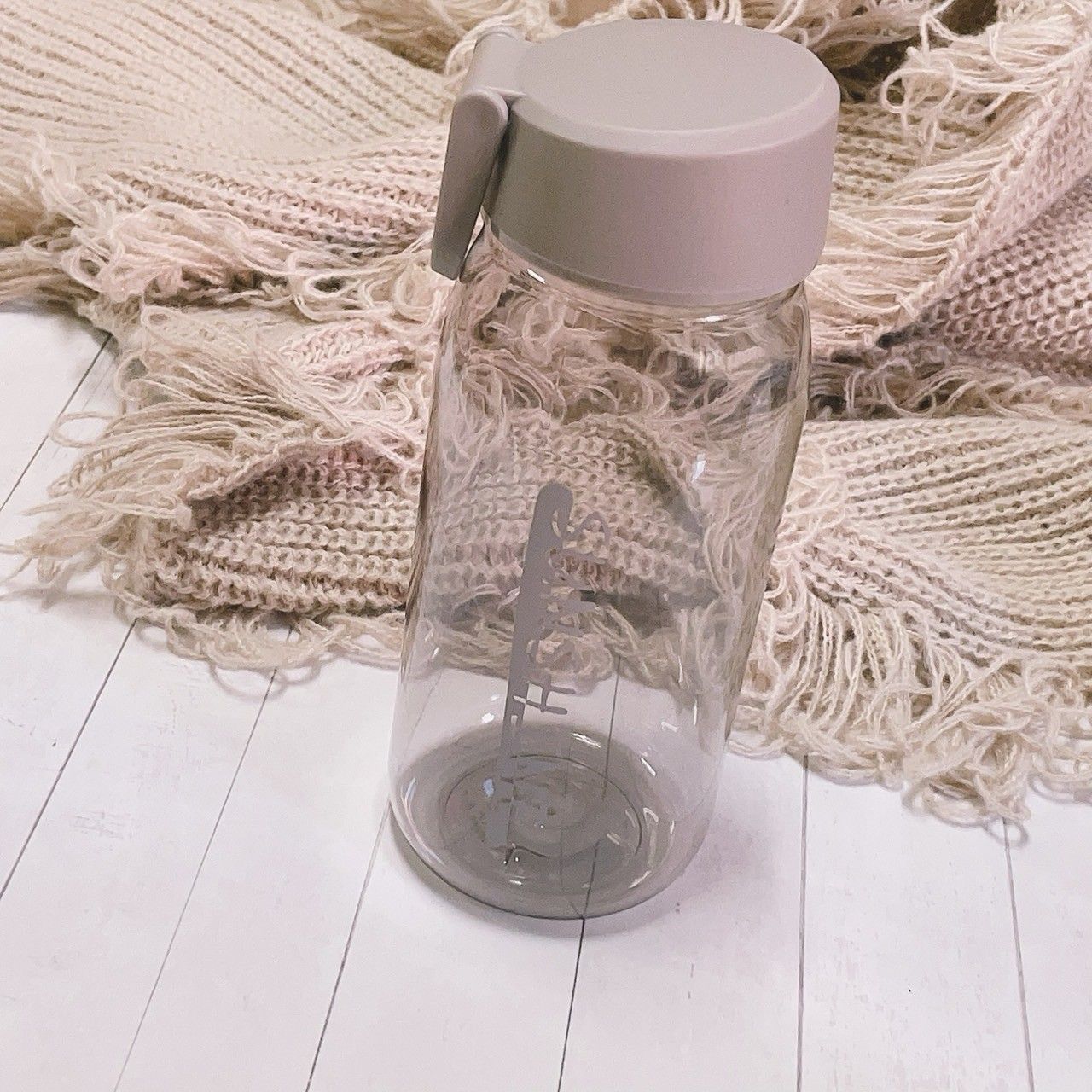 一個透明塑膠水瓶，配有灰色瓶蓋，放在白色木質表面上，旁邊是一塊帶流蘇的米色布料。
