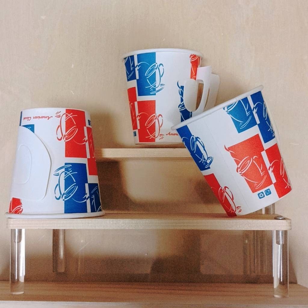 三個印有紅色和藍色方塊圖案以及白色茶壺的紙杯，擺放在多層木製展示架上。