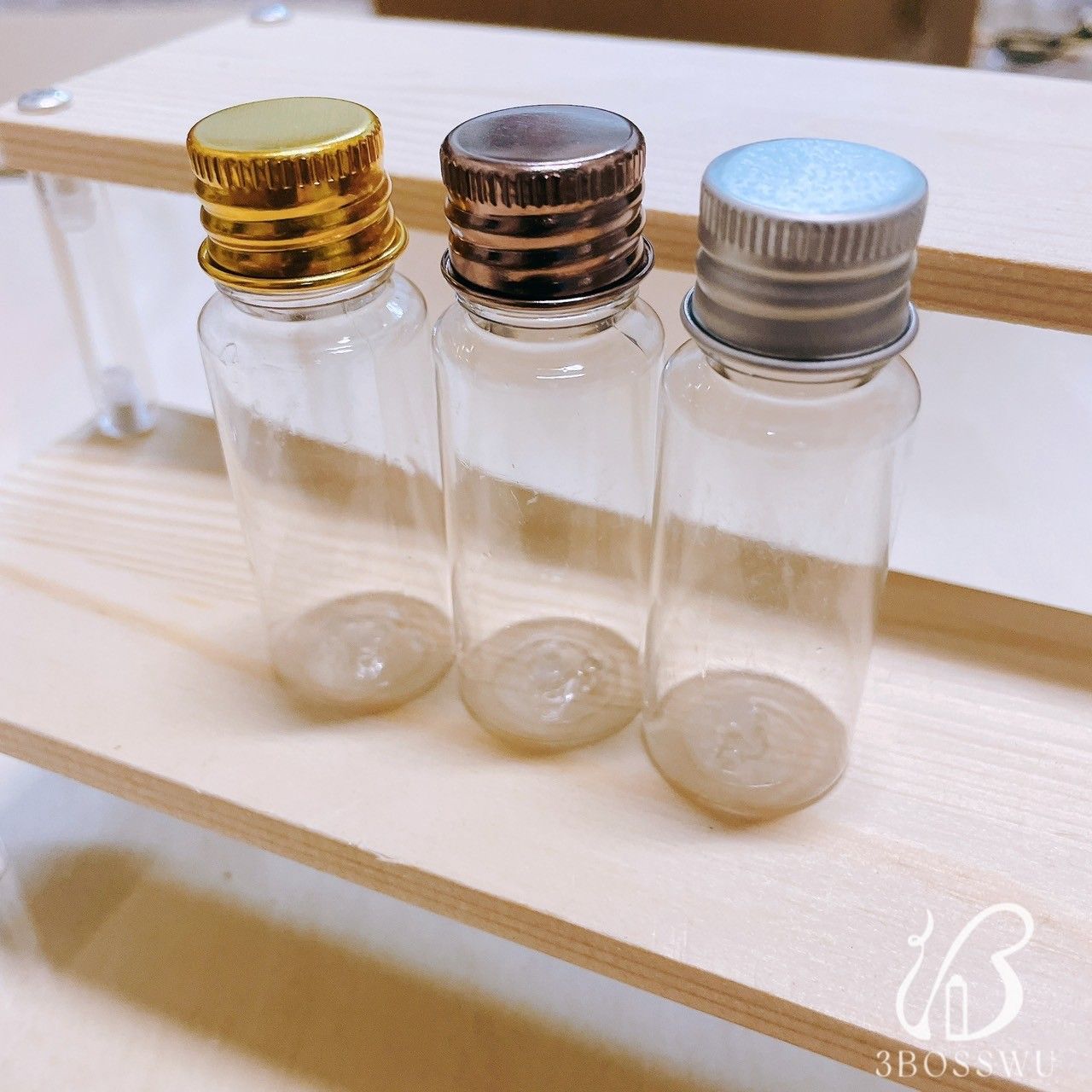 三個透明玻璃小瓶，分別配有金色、深色金屬色和銀色的螺旋蓋，擺放在一個淺色木製多層展示架上。