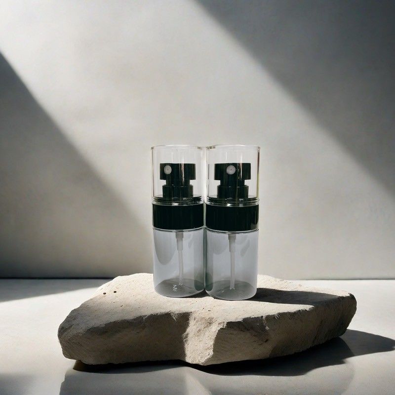 兩個空的透明玻璃噴霧瓶，配有黑色泵頭，並排放在一塊粗糙的淺色石頭上，靠著一面亮著燈的牆。