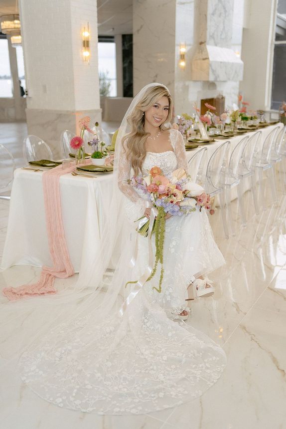A bride in a wedding dress is sitting at a long table holding a bouquet of flowers.