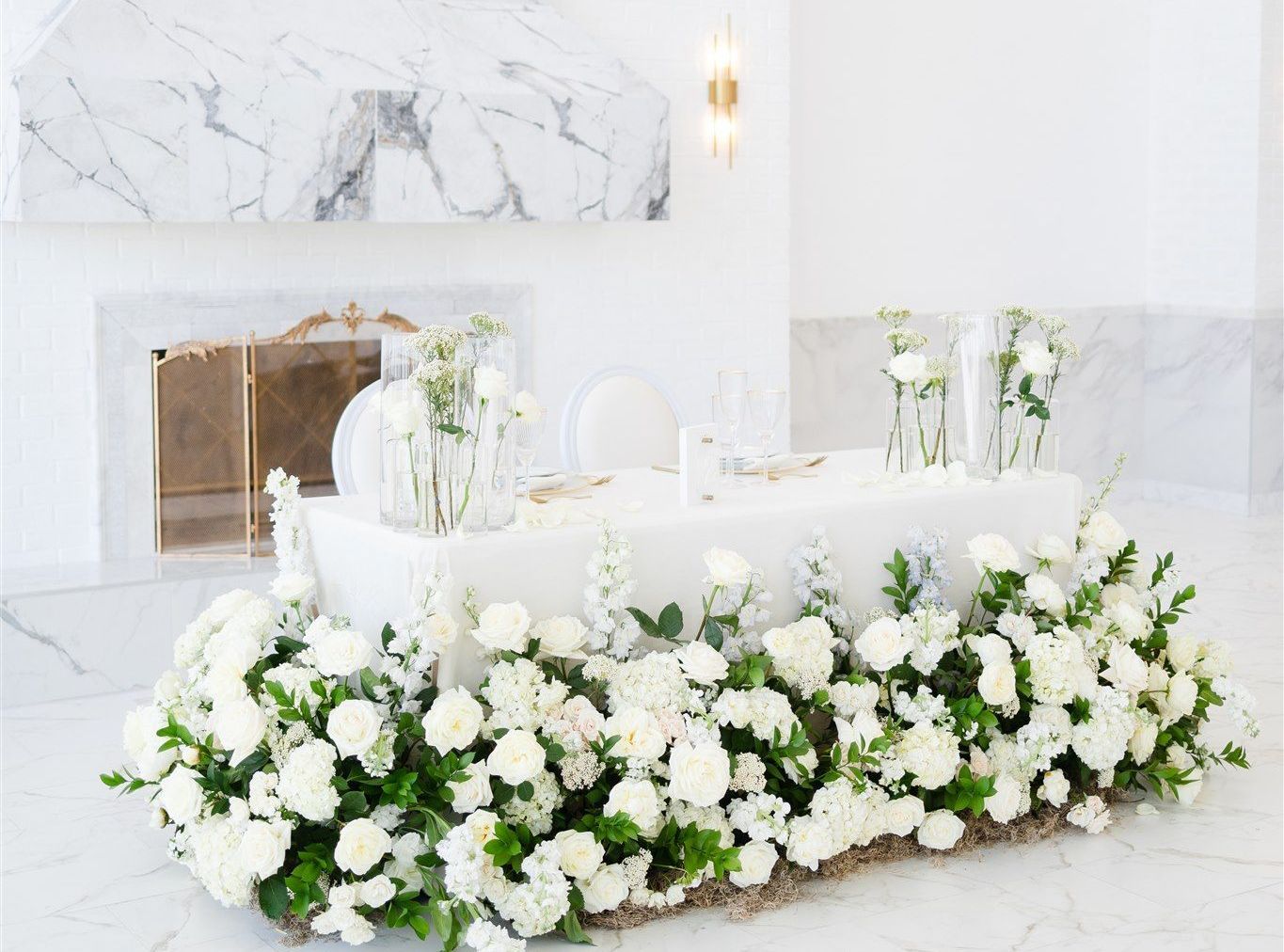 A long table decorated with white flowers and greenery in front of a fireplace.