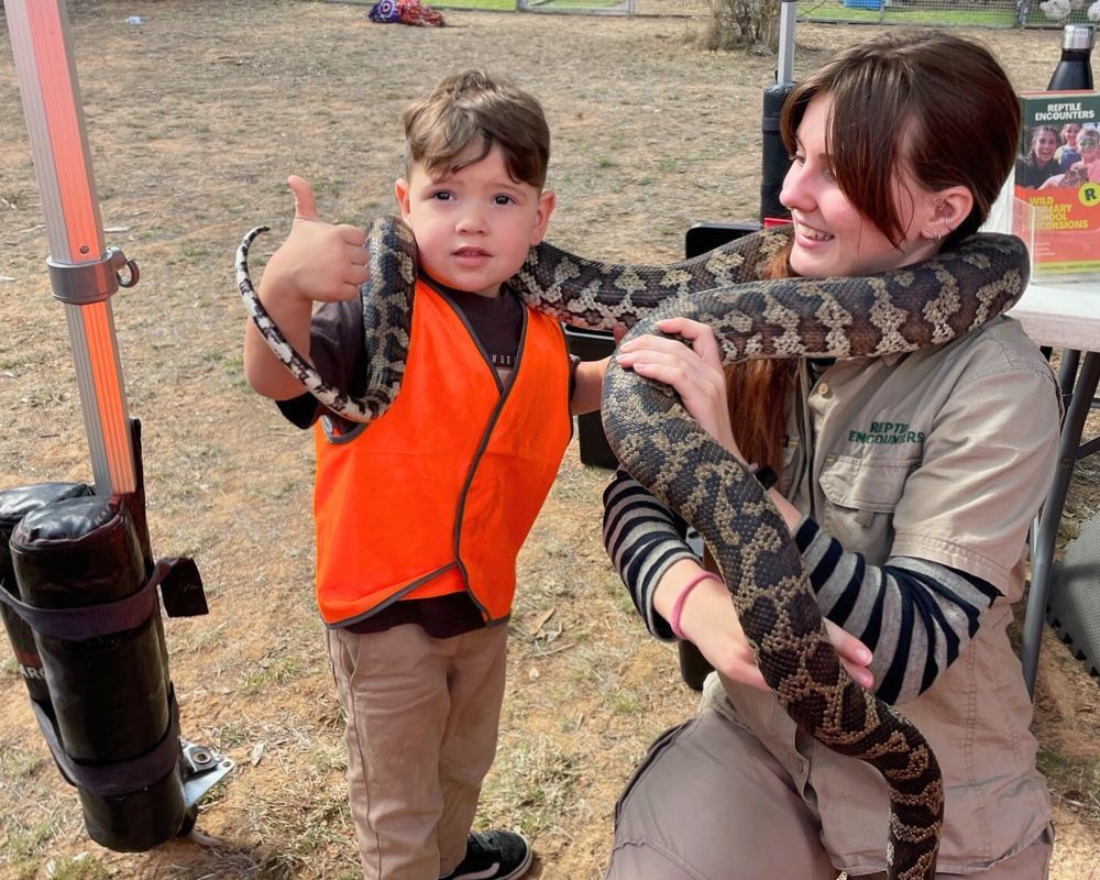 A young boy in an orange vest gives a thumbs-up, while a woman holds a large snake around their necks. Outdoor setting.