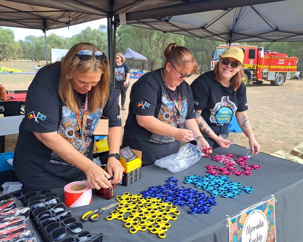Three women wearing matching t-shirts prepare colorful items at a table under a tent. They are smiling and working outdoors.
