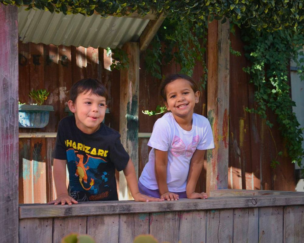 Two children smile, leaning out of a wooden playhouse window. The boy wears a black shirt; the girl wears a white shirt. The setting is outdoors.