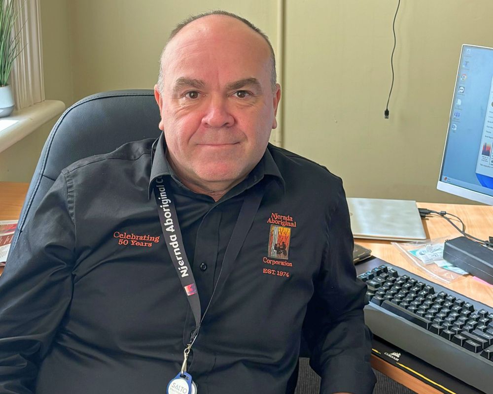 A man in a black shirt with text, seated at a desk with a computer and keyboard. He looks directly at the camera, in an office setting.