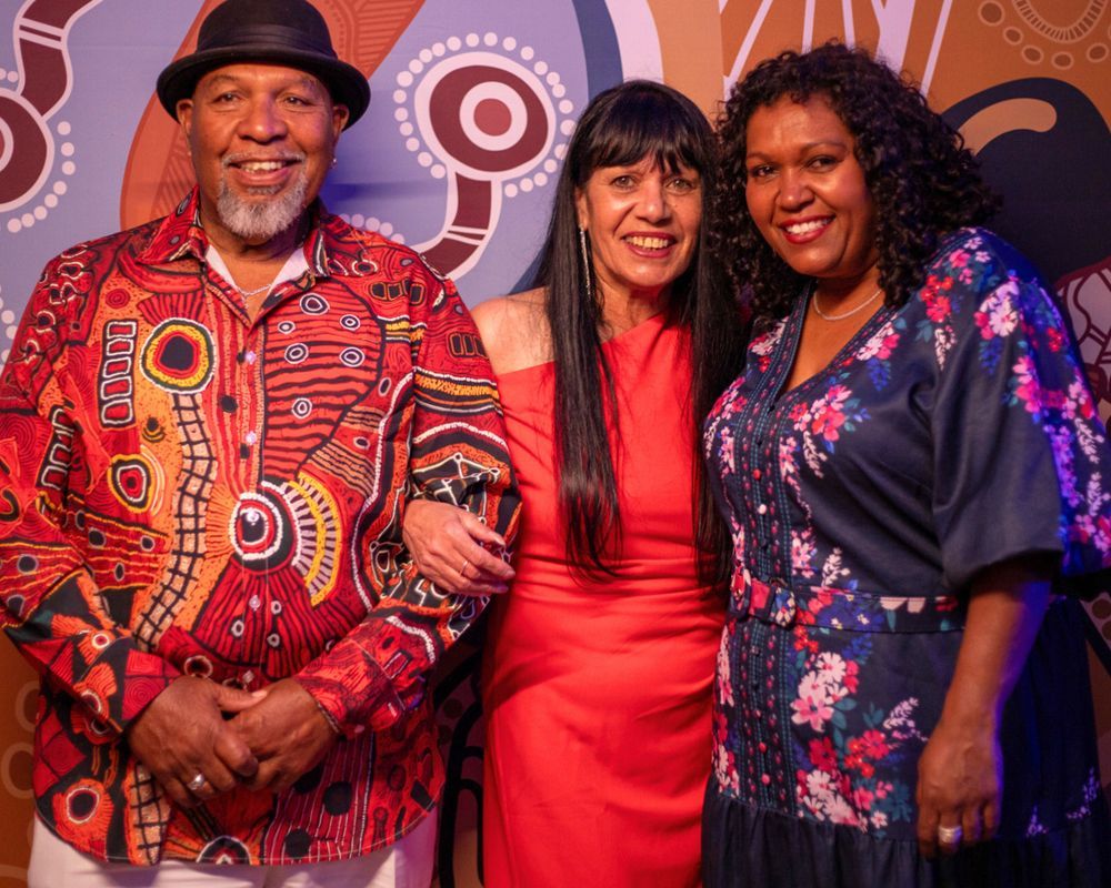 Three people posing in front of a backdrop with Aboriginal art. A man in a patterned red shirt and hat stands with two women in a red dress and a floral top.