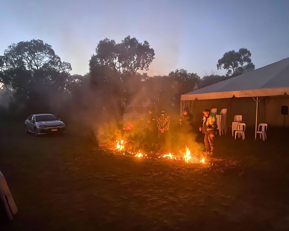 Firefighter battling a brush fire near a tent, a car, and trees. Evening light and smoke obscure the scene.