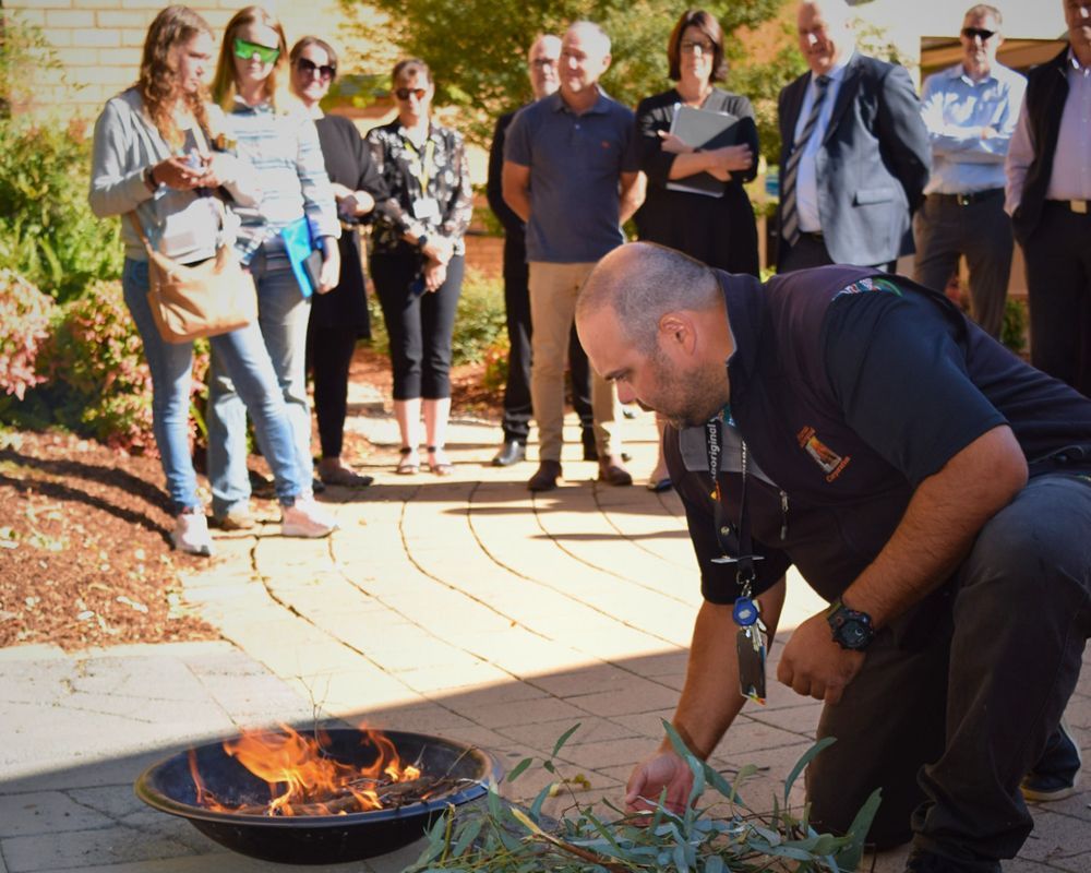 A man in uniform tends to a small fire in a bowl as a group of people watch outdoors.
