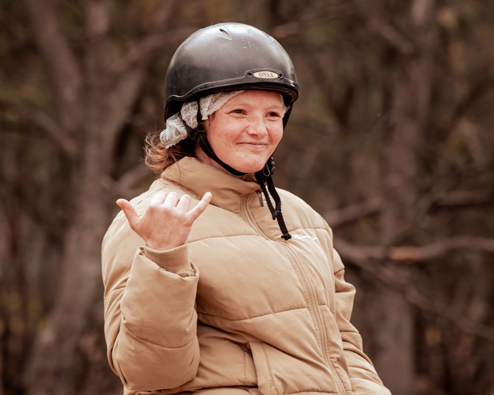 Young person wearing a riding helmet and tan puffer jacket, smiling and giving a shaka sign with their hand. Background is blurred trees.