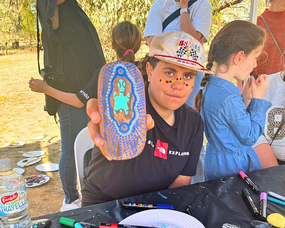 Young person with face paint holds up a painted rock with an Aboriginal design. They are seated outdoors, with other people visible.