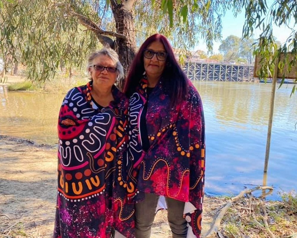 Two Indigenous women wrapped in patterned blankets stand by a river. One has grey hair, the other long red hair.
