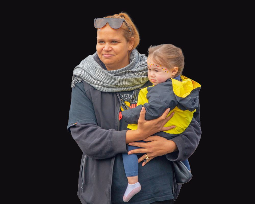 Woman with sunglasses holding a young child wearing a yellow jacket and face paint, on a black background.