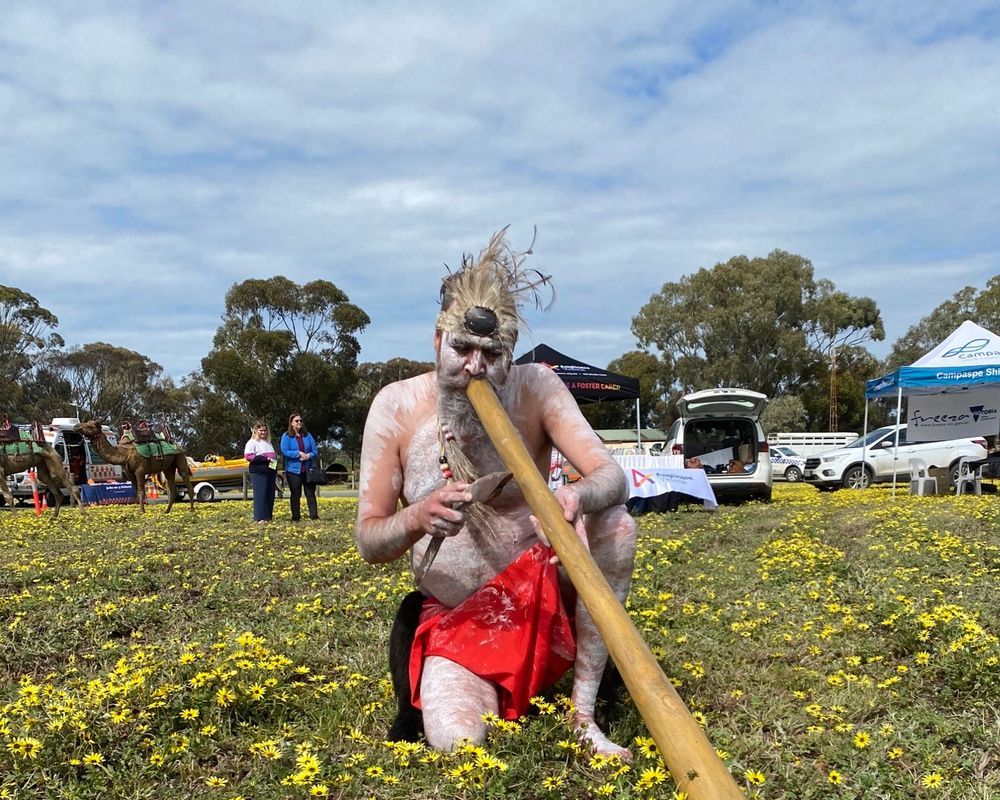 An Indigenous Australian man plays a didgeridoo in a field of yellow flowers under a cloudy sky. He is wearing traditional paint and a red cloth.