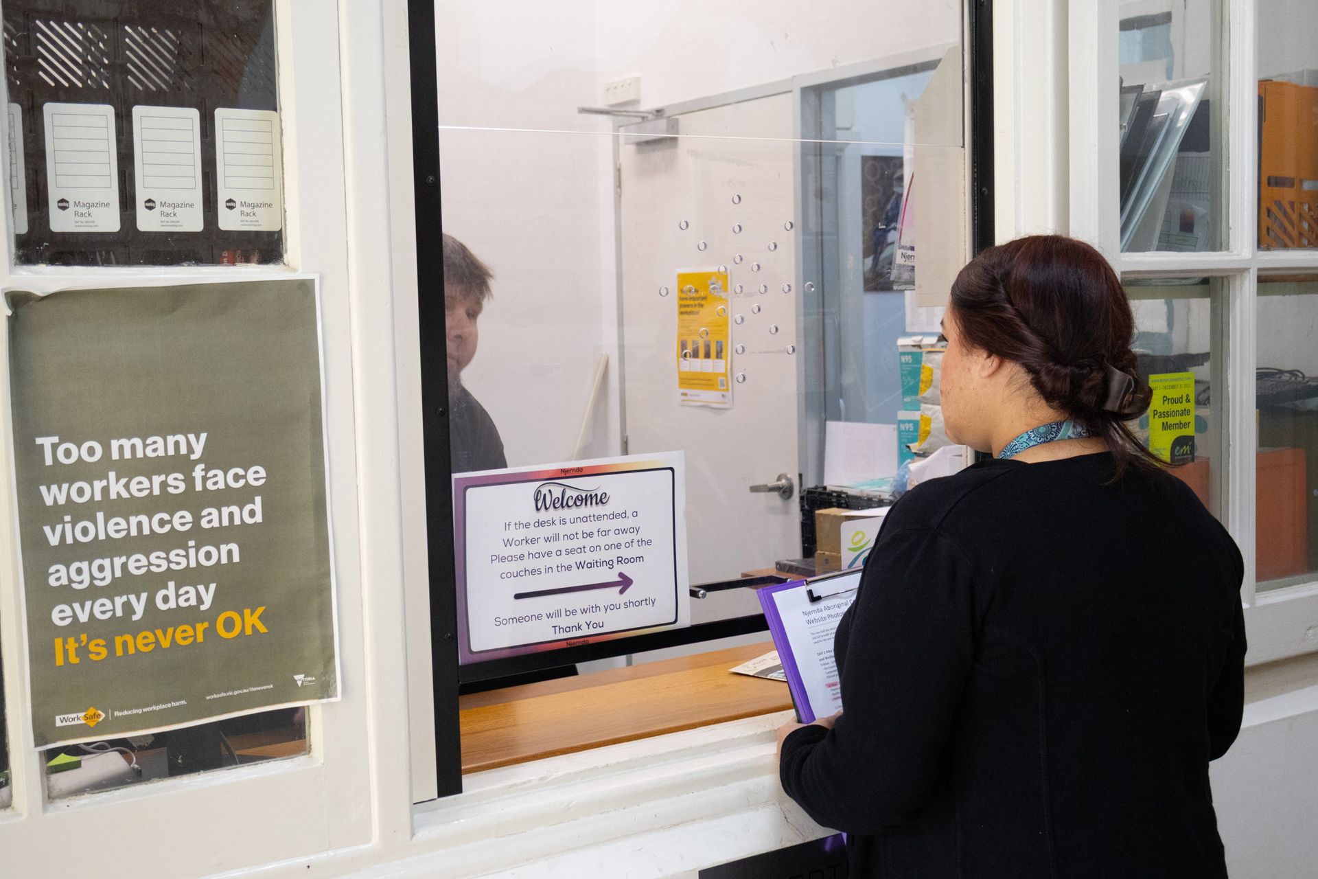A woman stands at a service counter with a sign about workplace violence. A person is visible behind the counter.