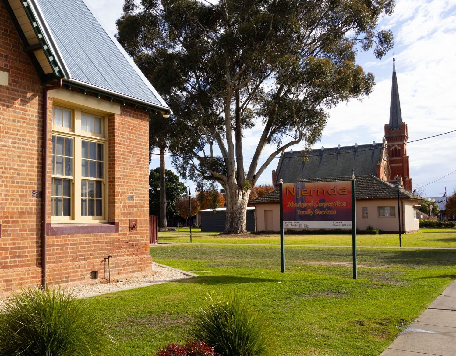 A brick building and a church with a steeple on a grassy lawn in a sunny setting. A sign stands in the foreground.