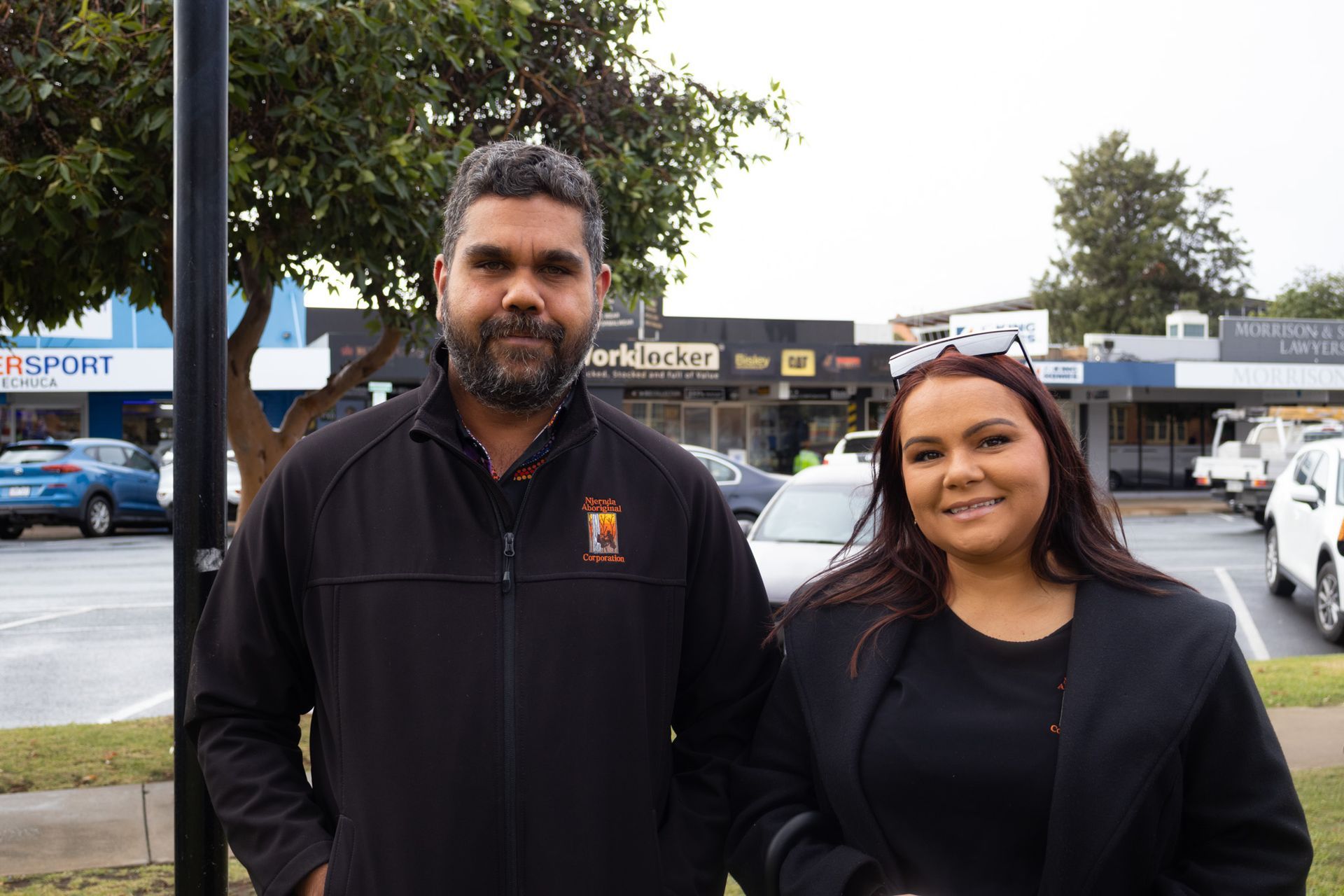 A man and woman stand outdoors in front of a strip mall. The man has a beard and is wearing a black jacket. The woman is wearing a black blazer and smiling.