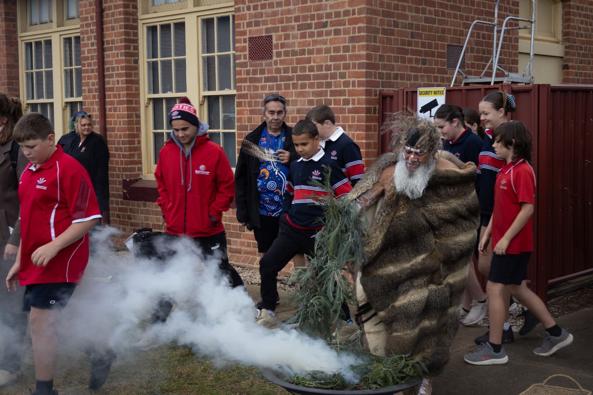 Children and adults watch a smoking ceremony outside a brick building. Smoke rises from a metal vessel with foliage.