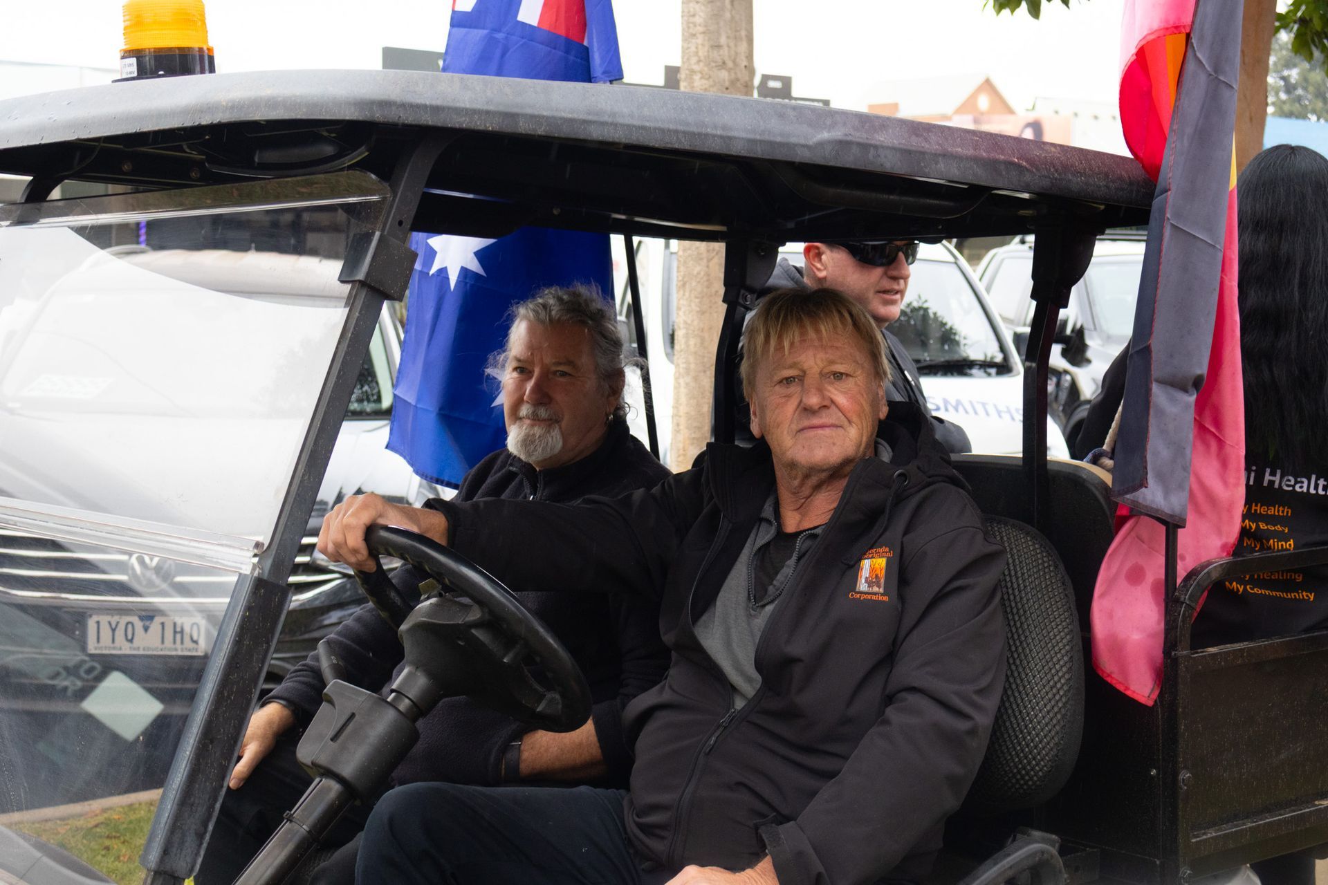 Two people in a golf cart, one driving, with Australian flags attached. They are outdoors, possibly at an event.