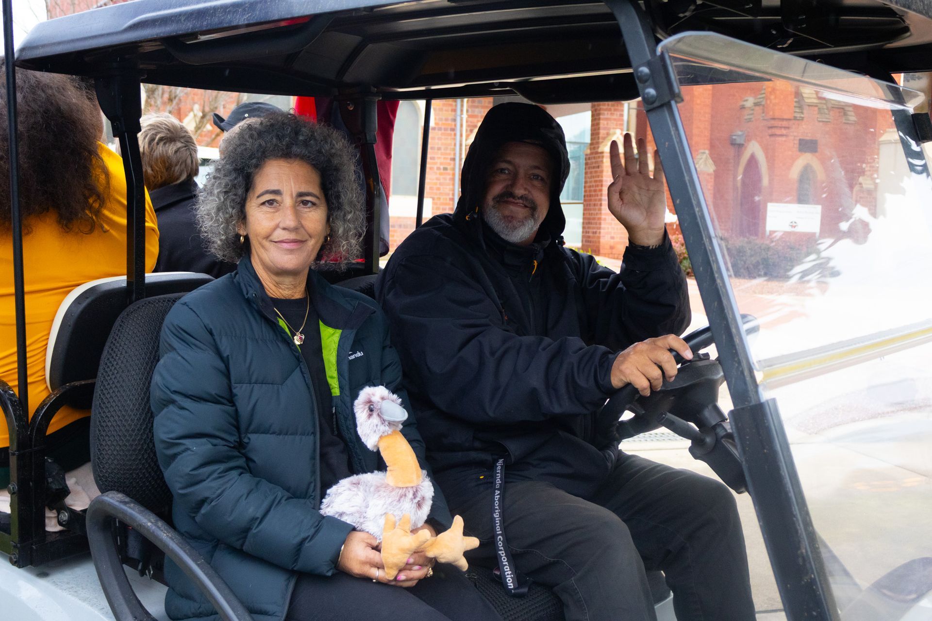 A man waves from a golf cart while driving, with a woman beside him holding a stuffed bird; a church is in the background.