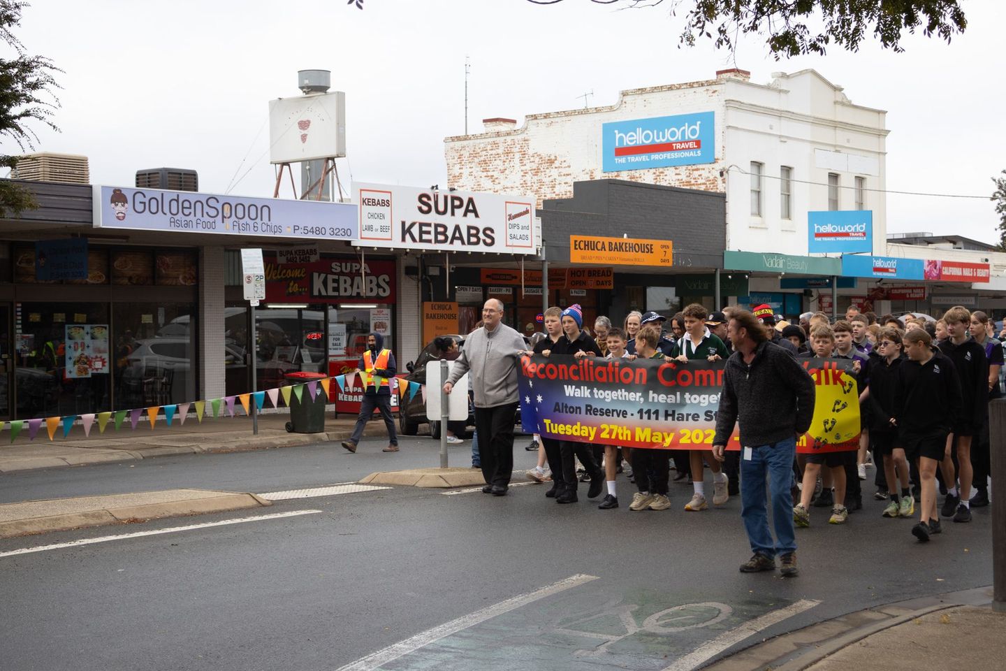 People march down a street holding a banner, with storefronts lining the road on a cloudy day.