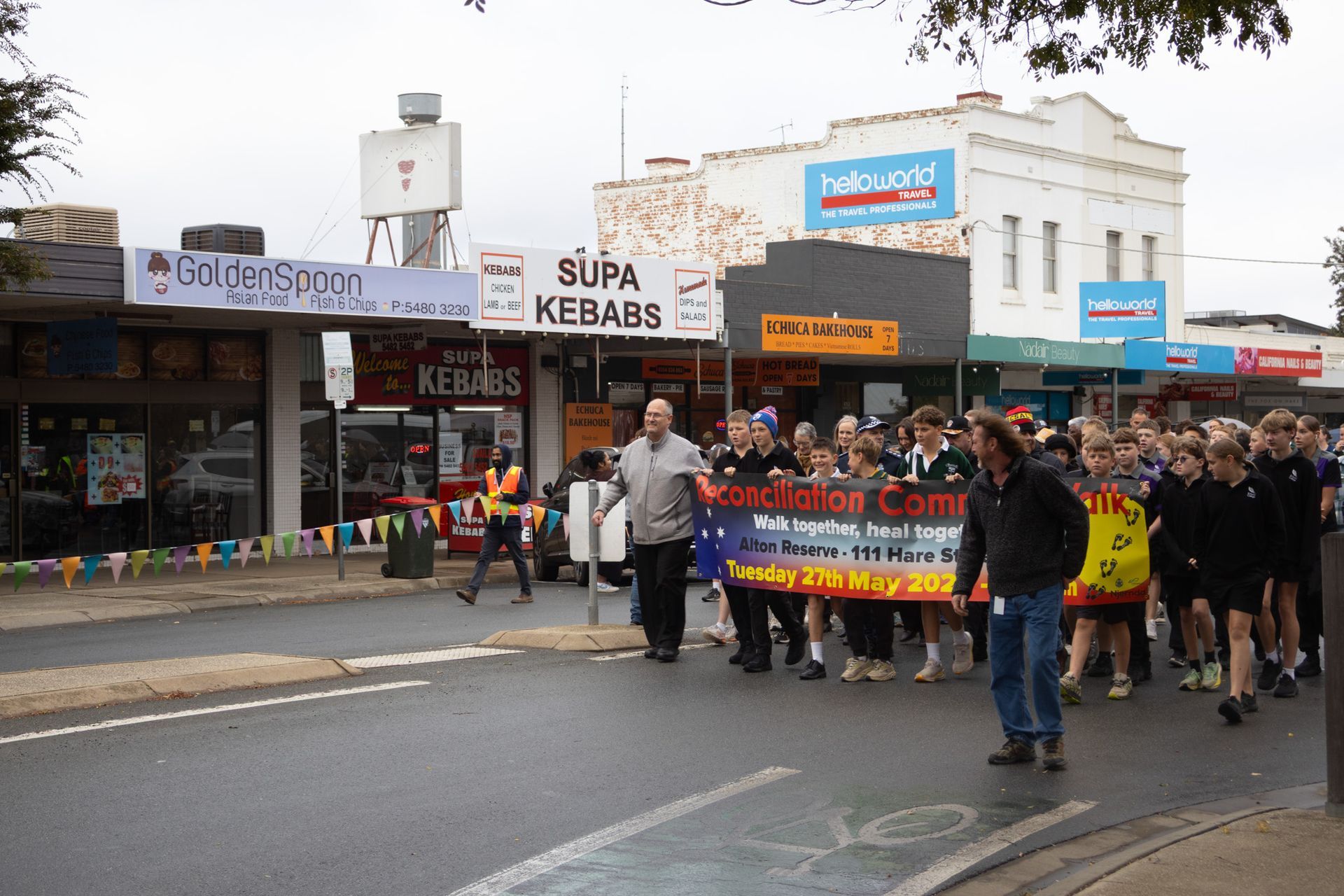 People march down a street holding a banner, with storefronts lining the road on a cloudy day.