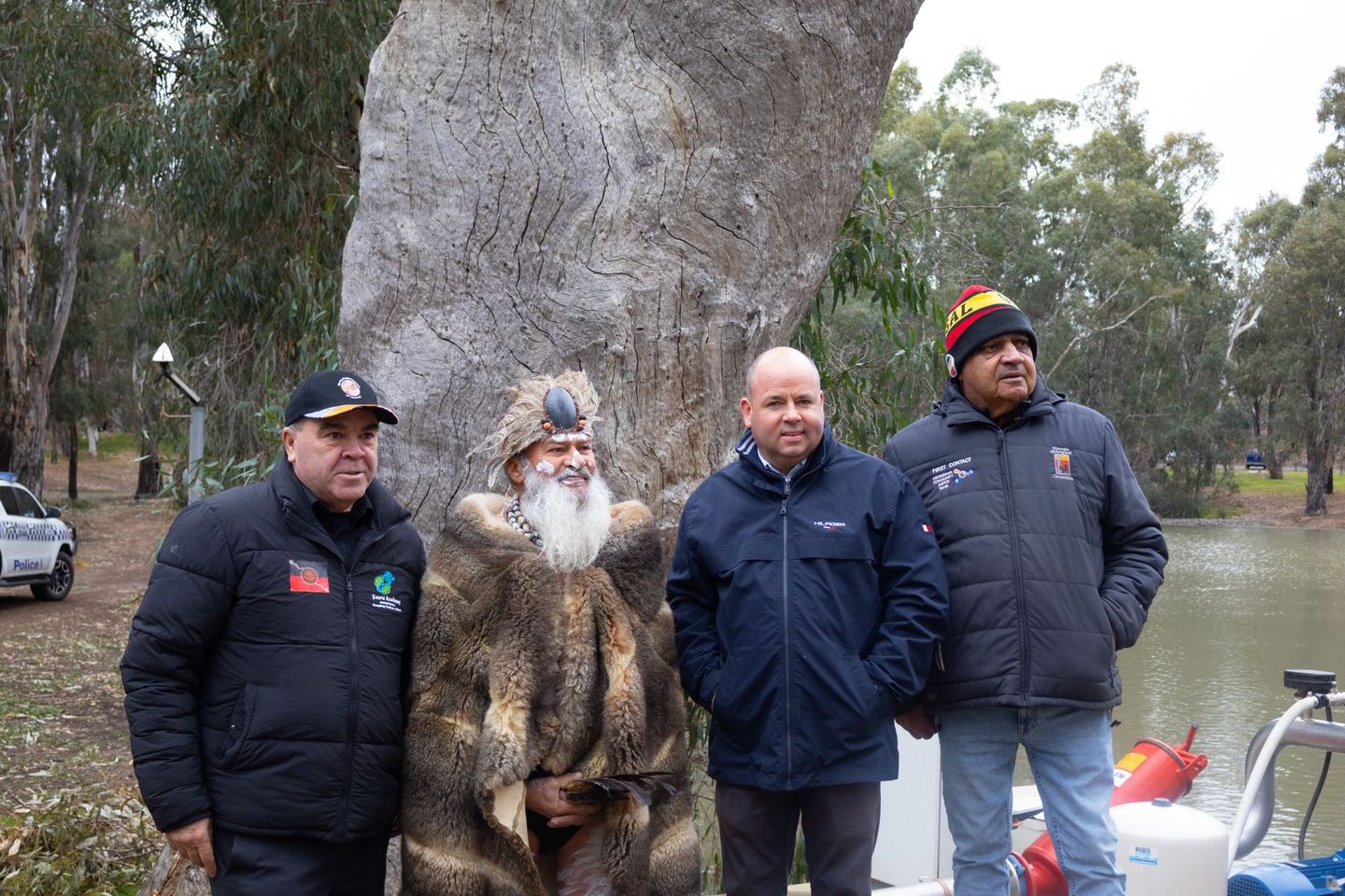 Four men stand near a large tree by water. One is in a fur coat and beard, others wear jackets and hats.