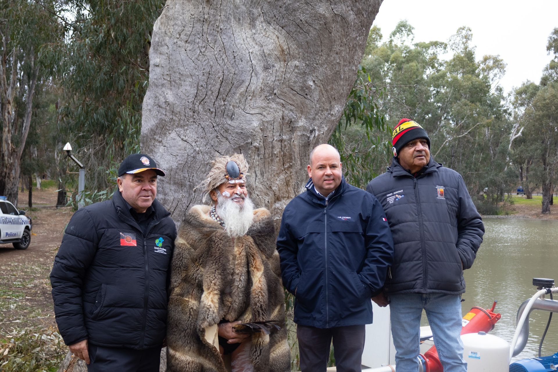 Four men stand near a large tree by water. One is in a fur coat and beard, others wear jackets and hats.
