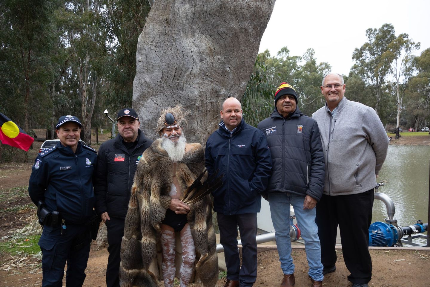 A group of six people pose outdoors by a large tree and water. One wears a traditional feathered garment.
