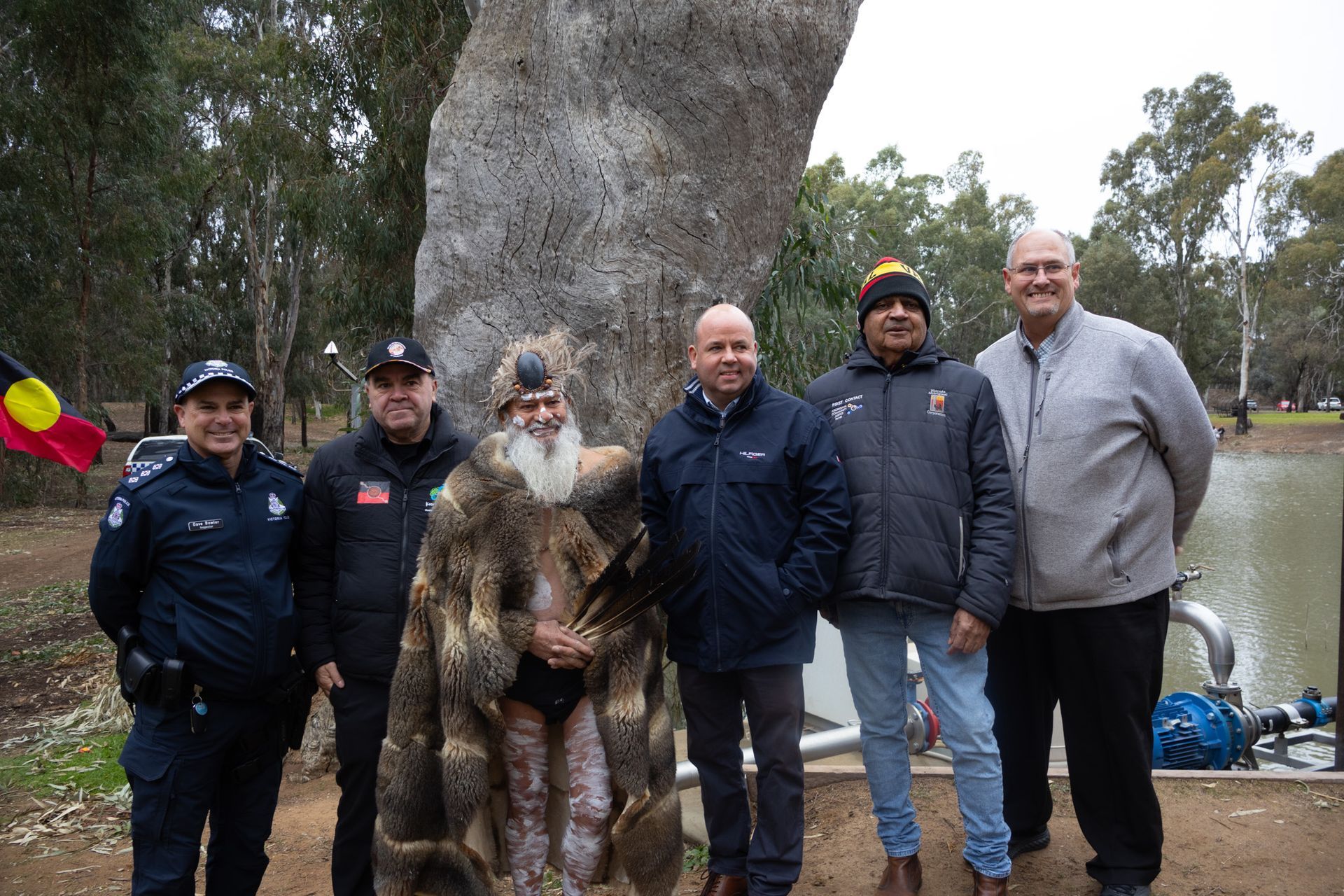 A group of six people pose outdoors by a large tree and water. One wears a traditional feathered garment.