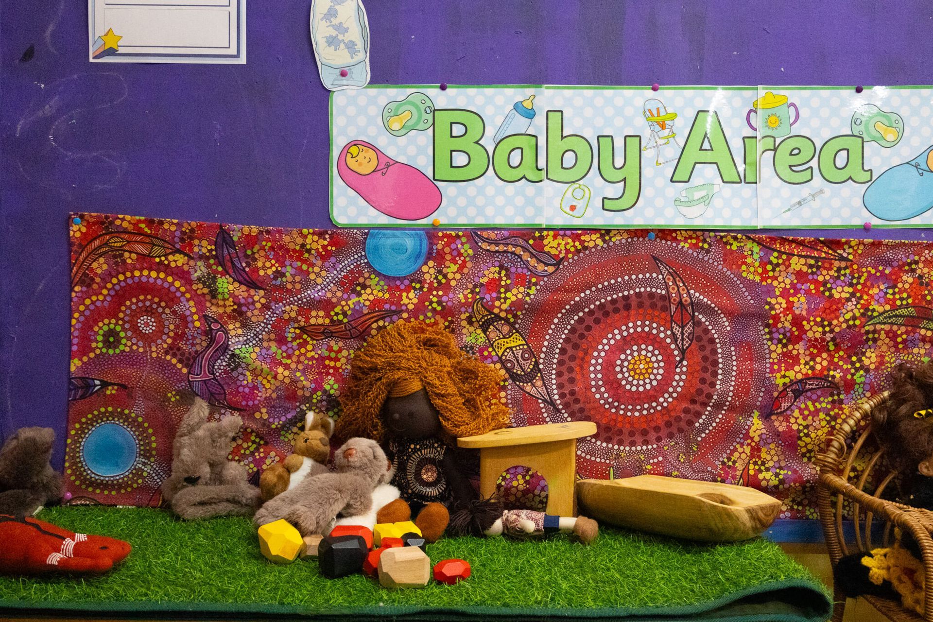 Baby area with a sign, colorful abstract background, toys on fake grass, and purple wall.