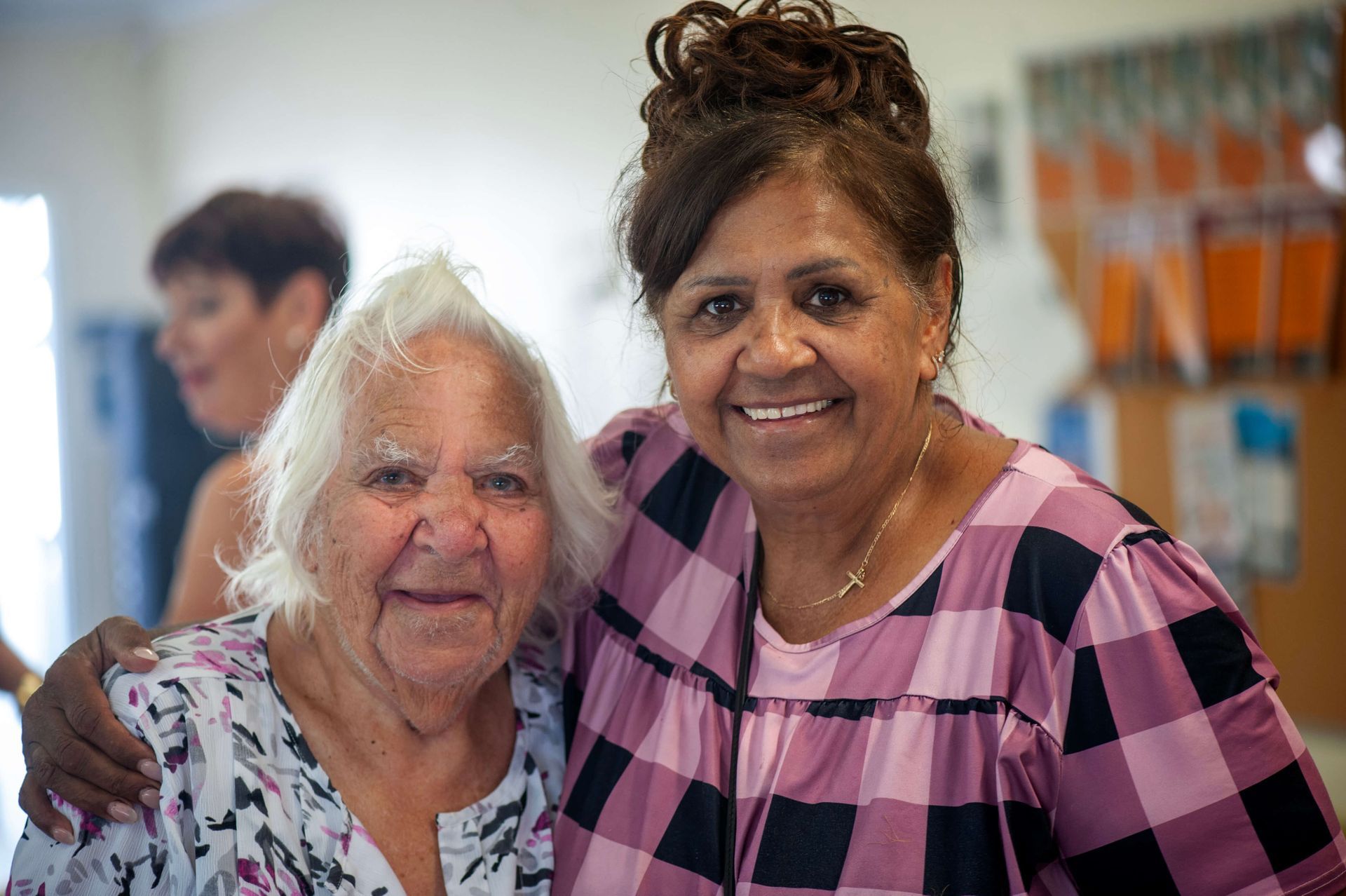 Two women smiling, one elderly with white hair, the other middle-aged with dark hair, indoors.