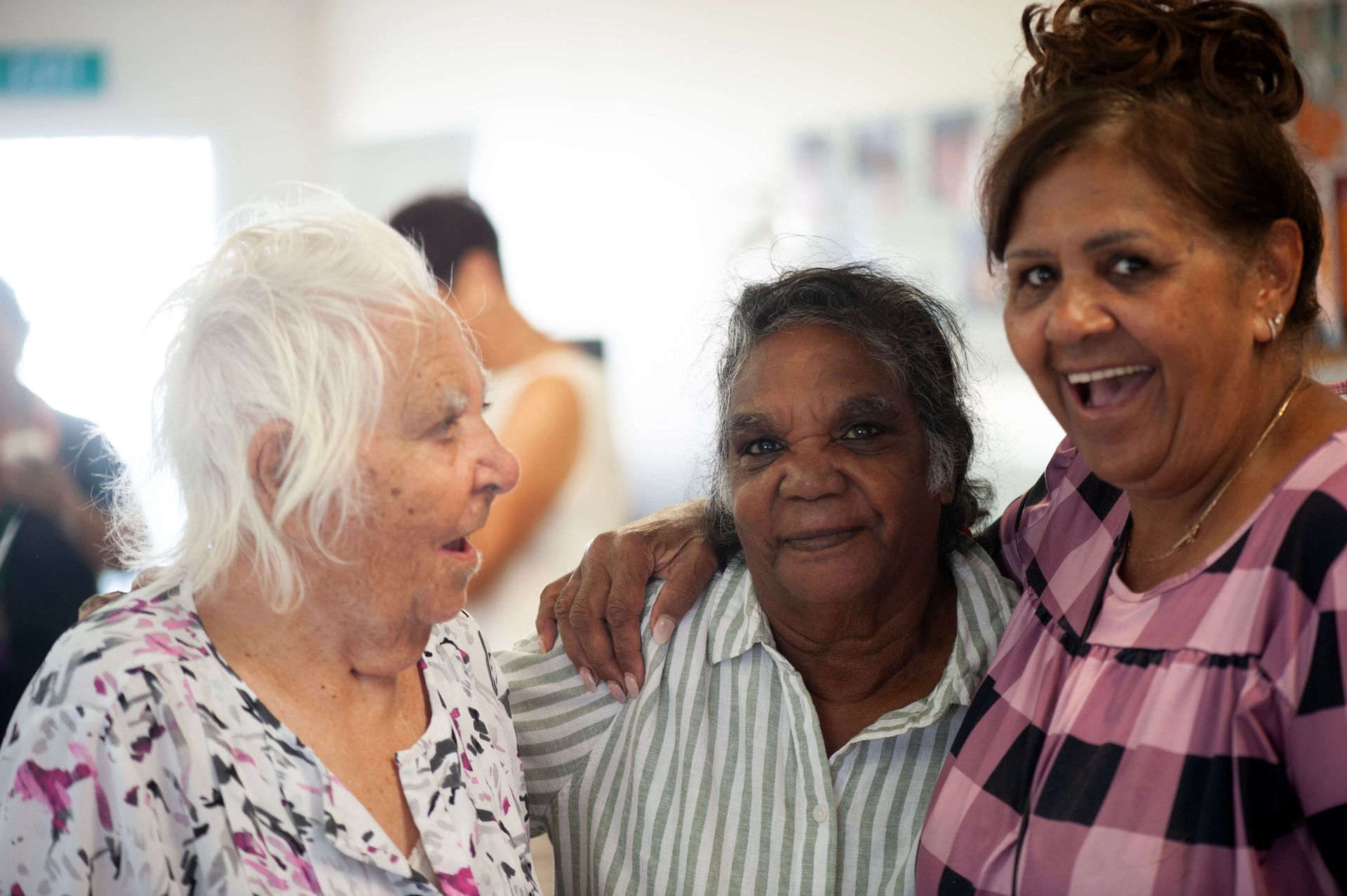 Three smiling elderly women, one with white hair, arm in arm indoors; one has her arm around the other, with a pink patterned top, the others have neutral expressions.