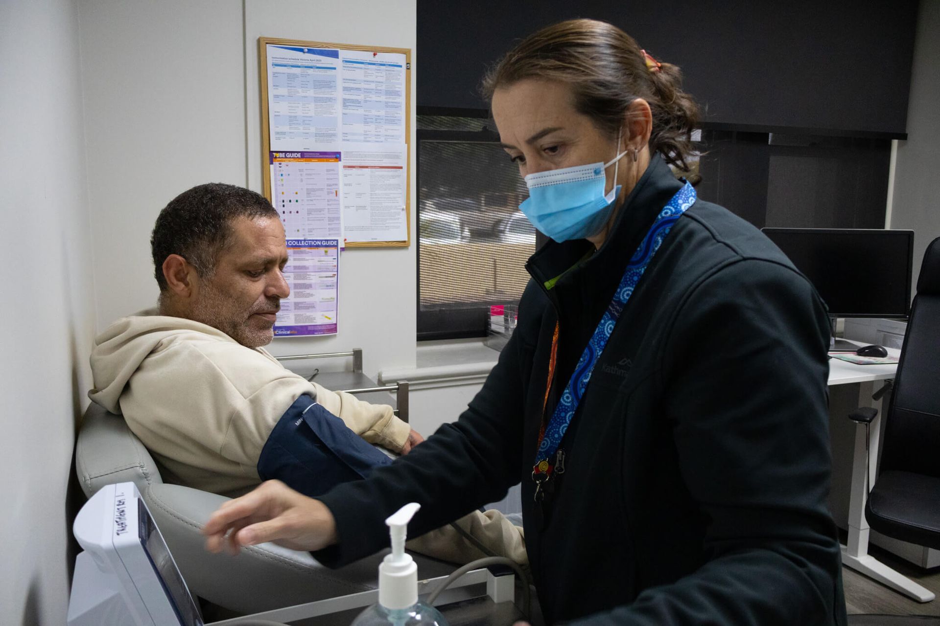 A health worker in a mask takes a man's blood pressure. They are in a medical office.
