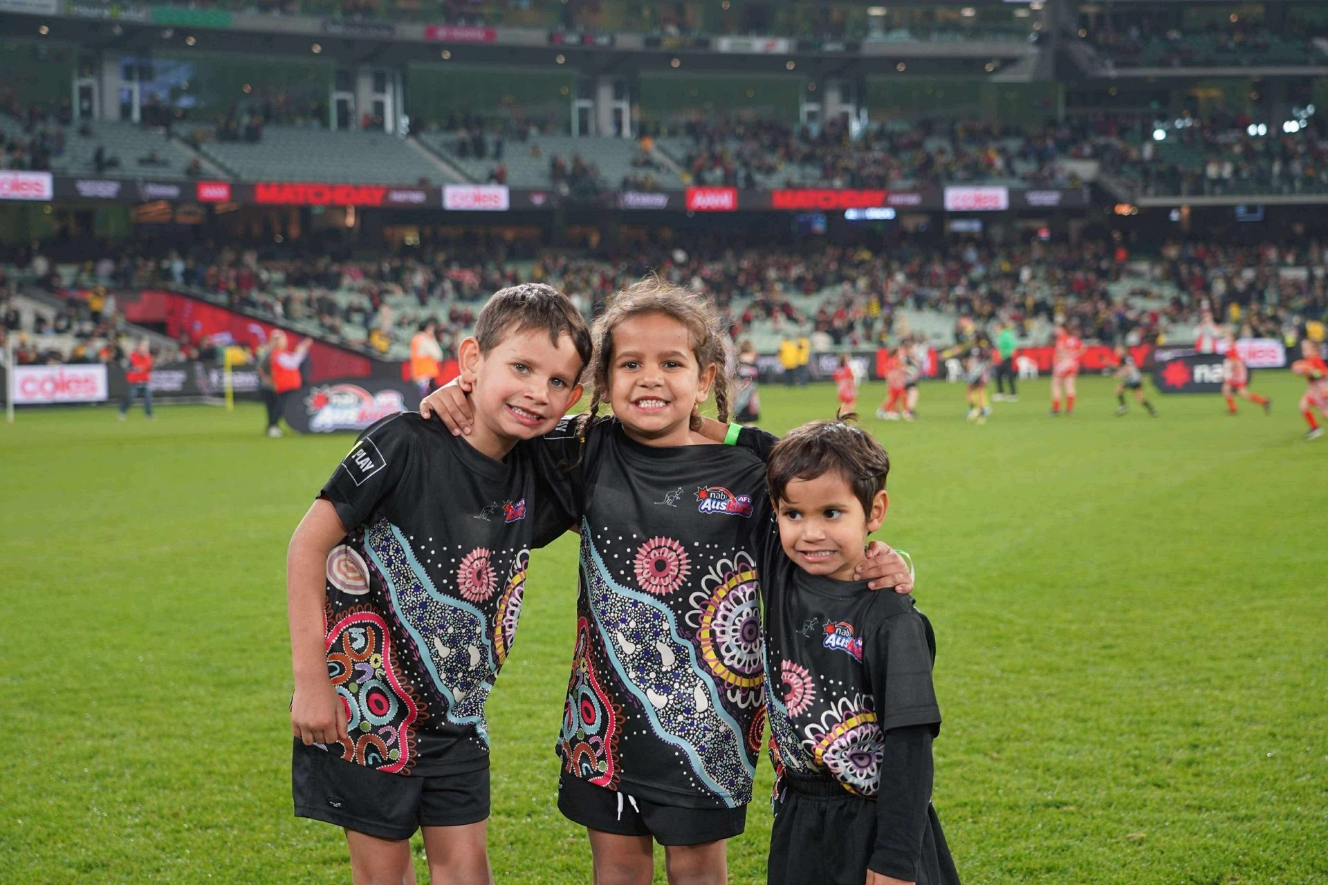 Three young Indigenous children smiling on a sports field, wearing matching black jerseys with colorful artwork, with a stadium crowd in the background.
