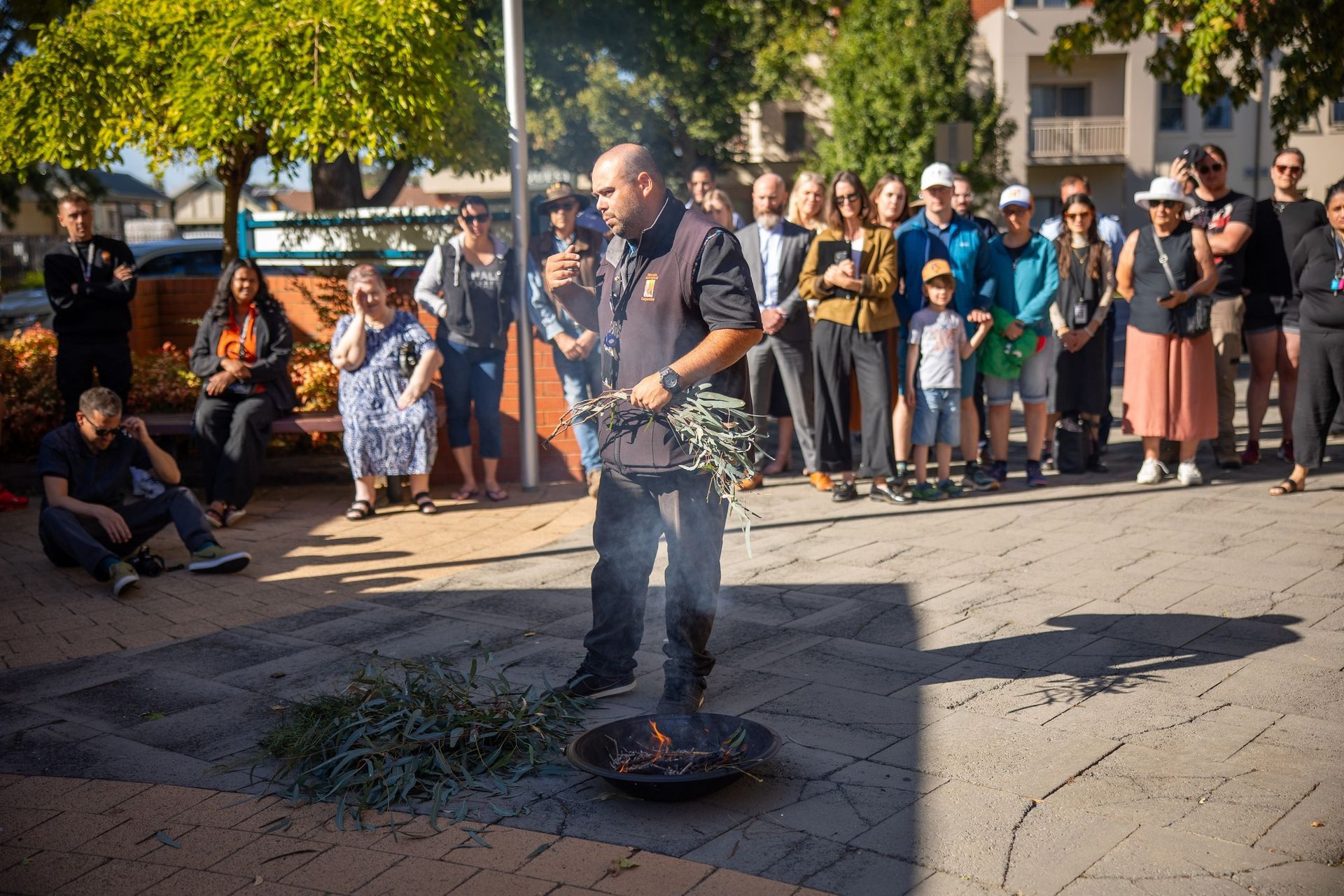 Man performing a smoking ceremony with burning leaves, surrounded by onlookers in an outdoor setting.
