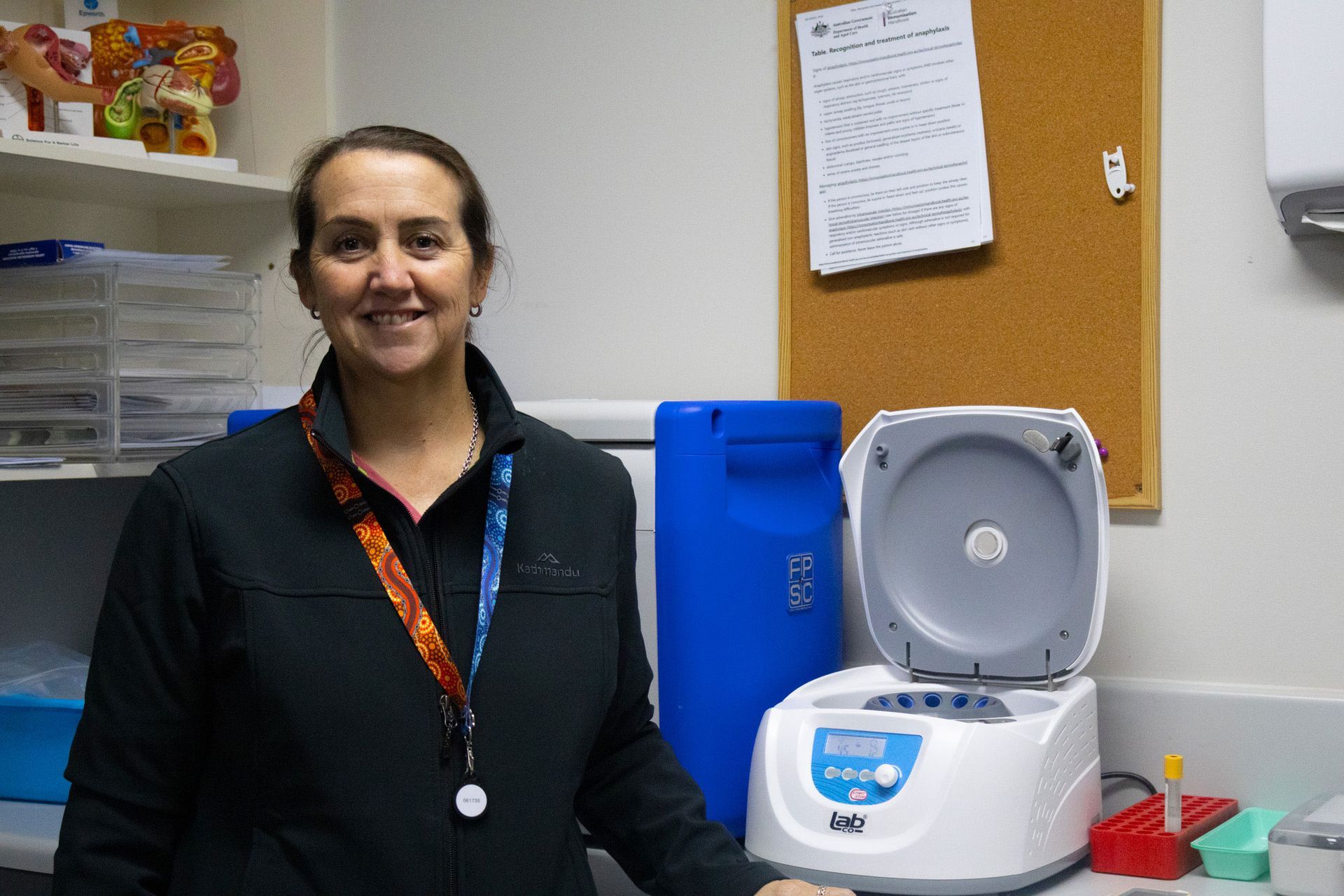 Woman in a lab coat smiles next to a centrifuge.  A blue drum and corkboard with notes are in the background.