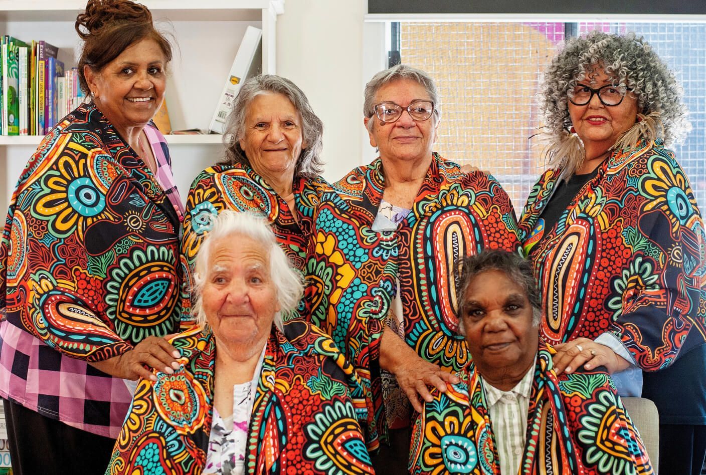 Six Indigenous women in colorful patterned jackets, smiling indoors.
