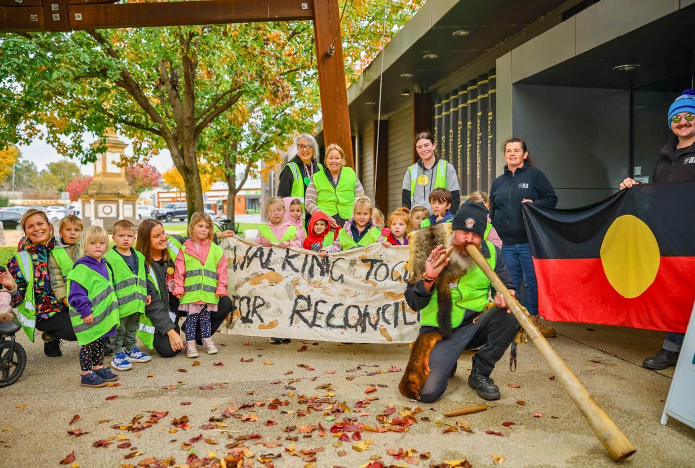 Children and adults in vests and a didgeridoo player gather outside a building with the Aboriginal flag and a banner that says 