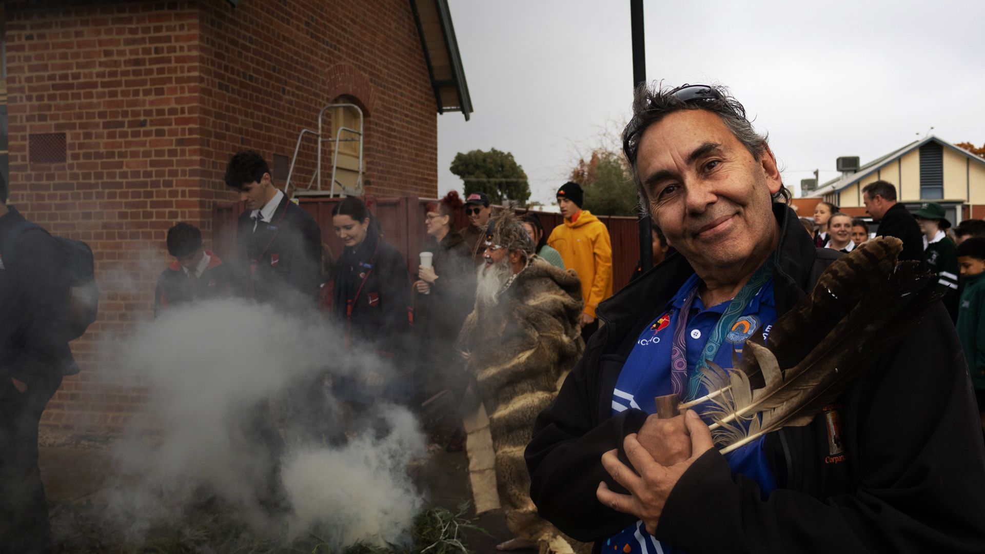 Person smiling, holding brass instrument, in front of smoking ceremonial fire, with students in background, outdoors.