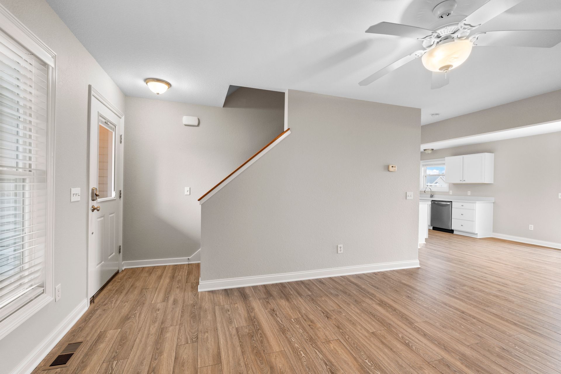 Interior view of a living space with wooden floors, staircase, and white kitchen in the background.