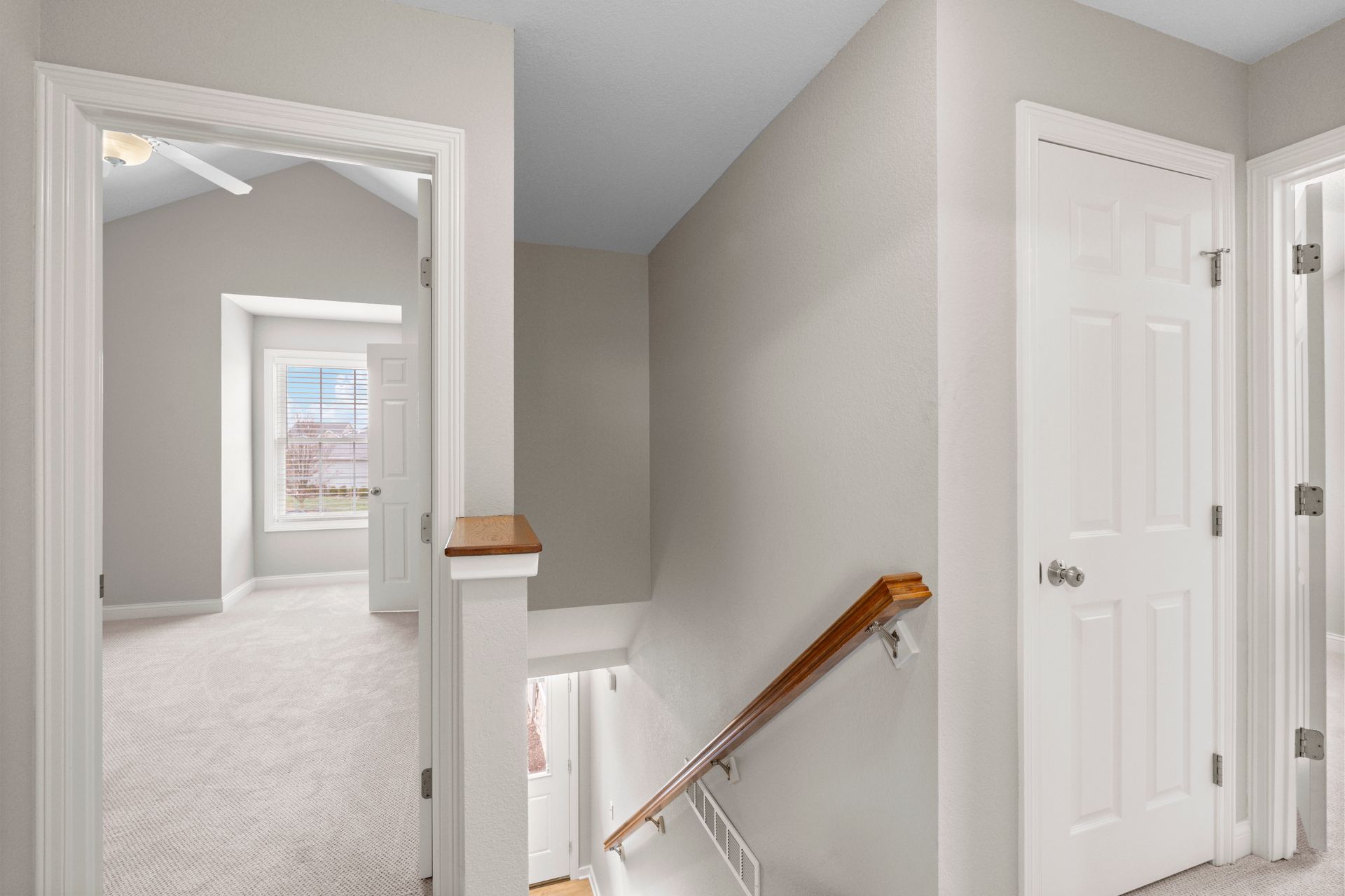 Hallway with doorway to a room with a window, white walls, carpet, staircase, and two white doors.